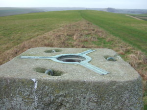 The top of the trig point, showing attachment fittings for a theodolite. In the blurry middle distance is the beacon, a metal basket atop a high pole.